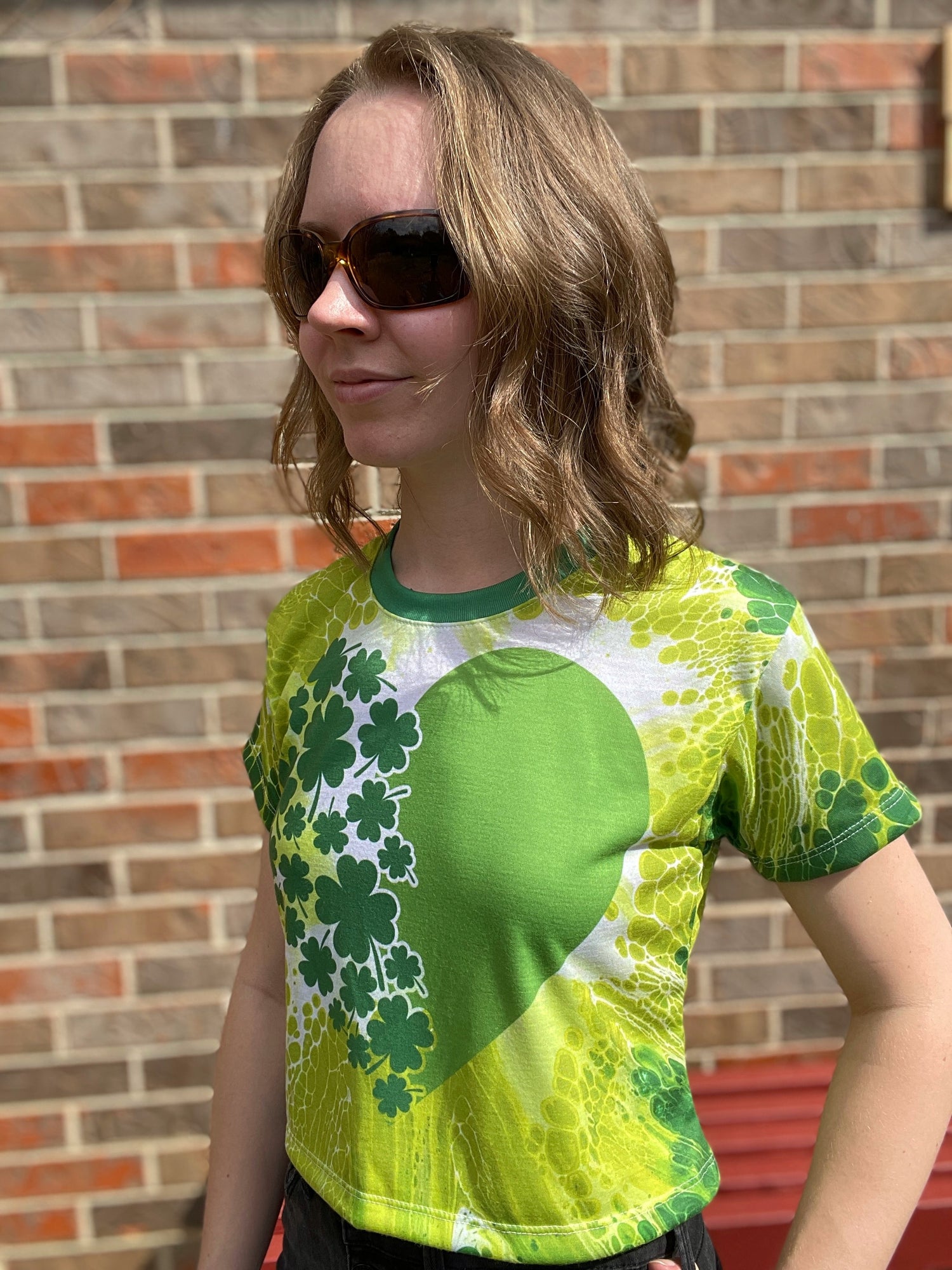 Artist KMKC stands in a Saint Patrick's Day themed t-shirt with green and white clovers, heart, and fluid art background.